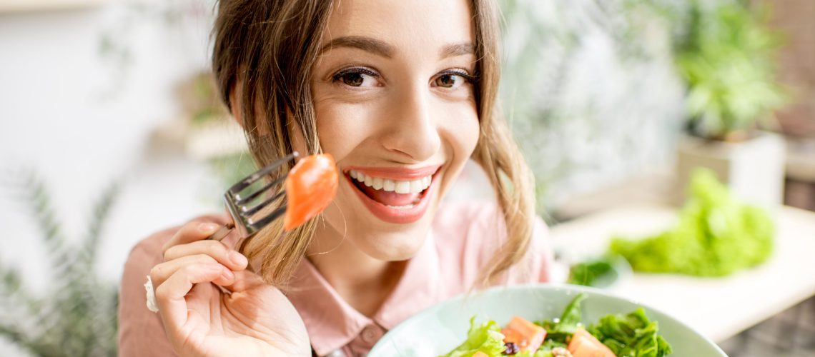 Young and playful woman eating healthy food sitting indoors on the green background